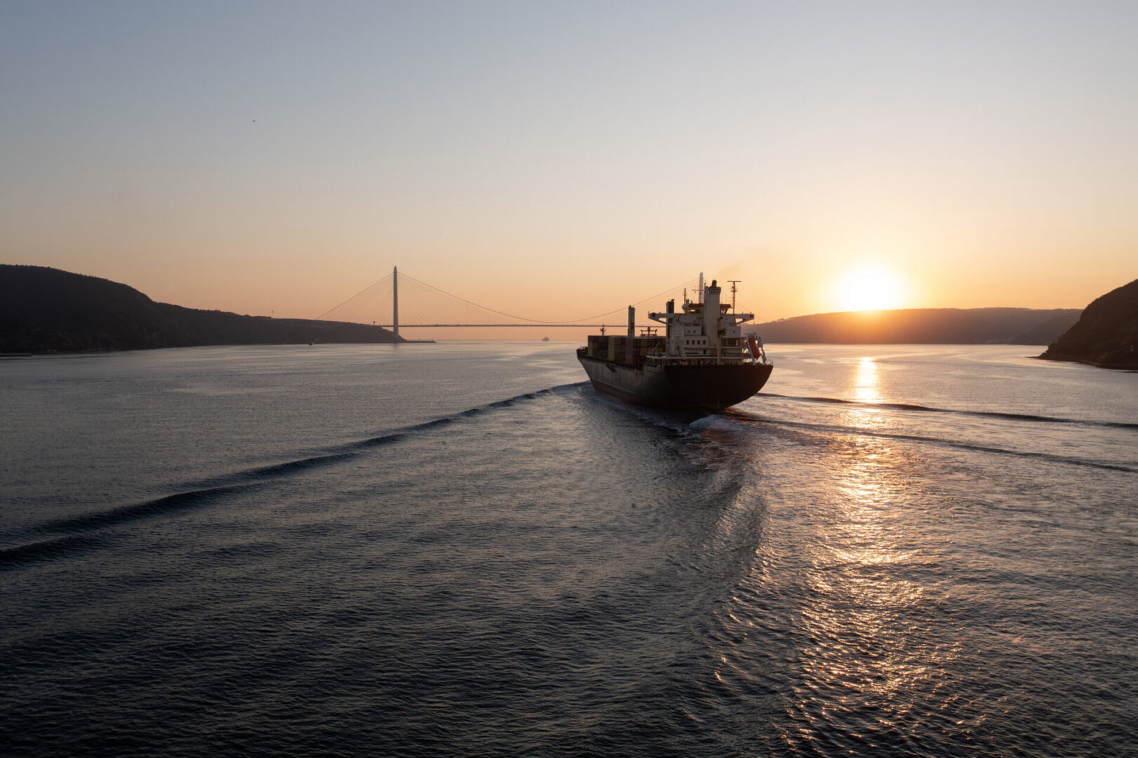 Cargo ship sailing toward sunset with bridge